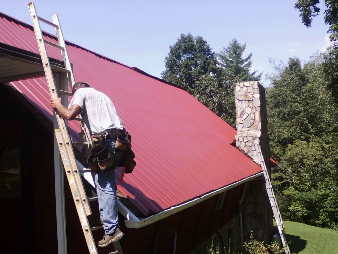Roofer on a ladder beside a red metal roof and stone chimney — Soffit & Fascia Repair by Roofing Brothers in Paramount